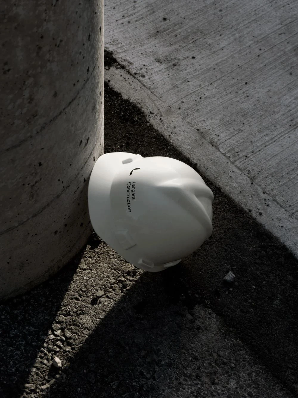 206d17e5f1d82c1d9dbb8b6bba8a6437 A white construction hard hat rests on the ground, leaning against a concrete pillar, on a surface of gravel and concrete.