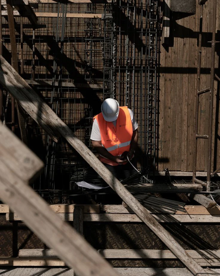 4b8ad3c75df60aacaae0bc41bb15b391 A construction worker in an orange safety vest and white helmet is inspecting steel reinforcement bars at a building site.