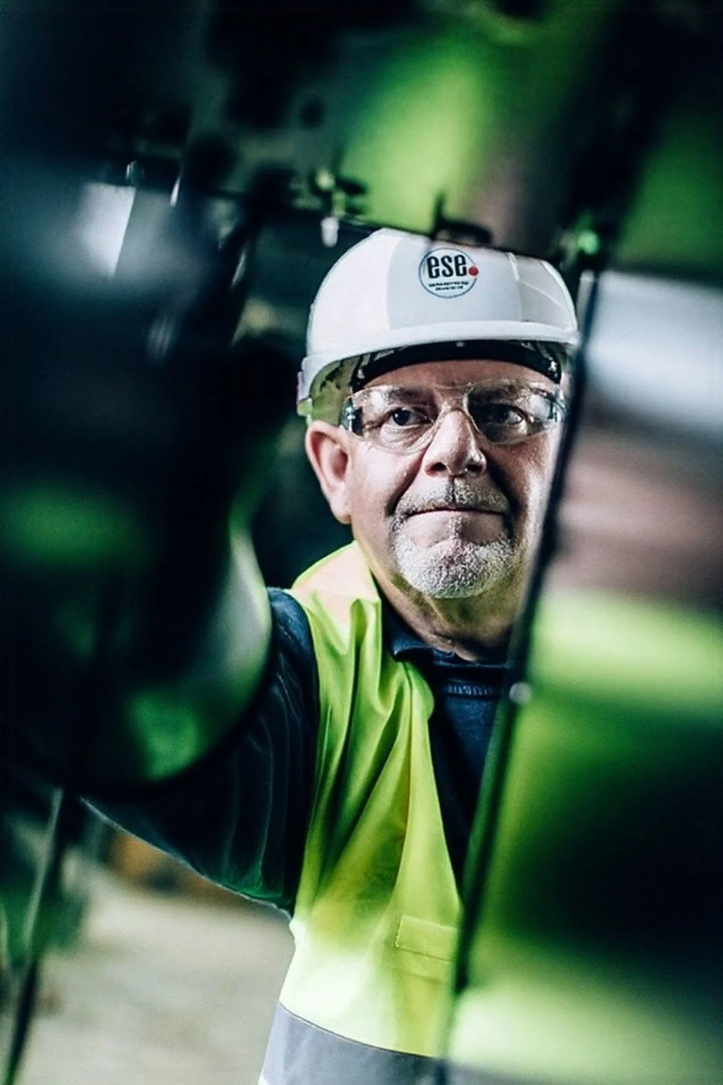 6ece82c331b5bfc8568ad84ada674aa0 An older man wearing a hard hat, glasses, and a high-visibility vest works with industrial machinery, viewed from a low angle.