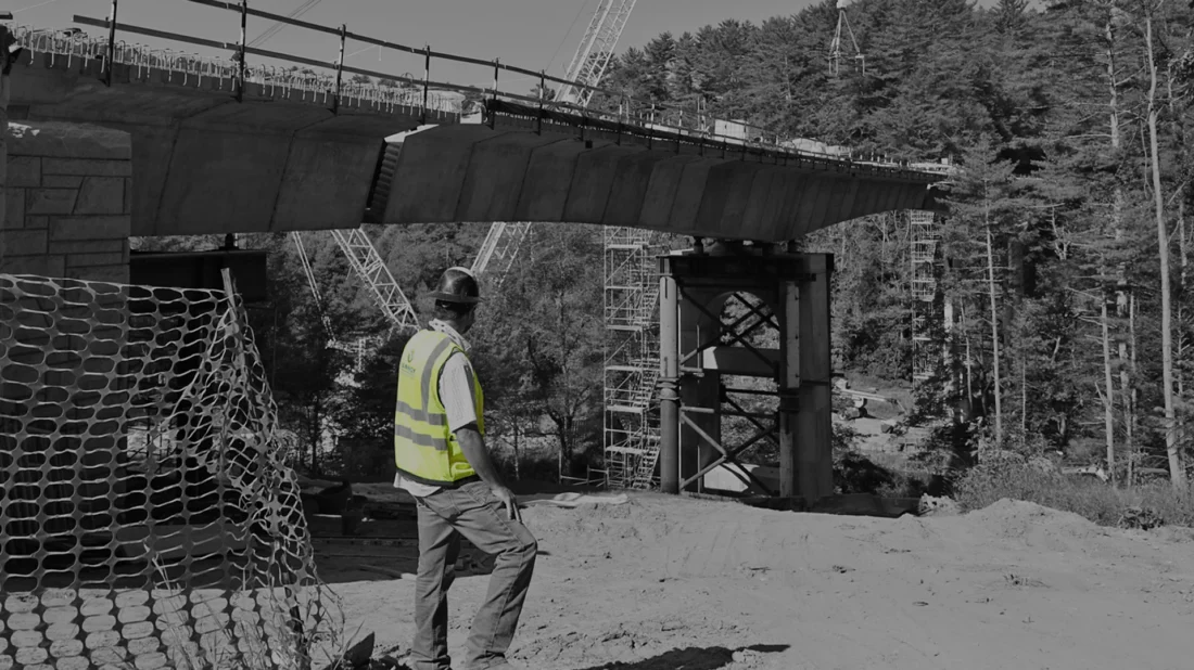 A construction worker in a safety vest stands near a partially built bridge, with cranes and scaffolding visible in the background.