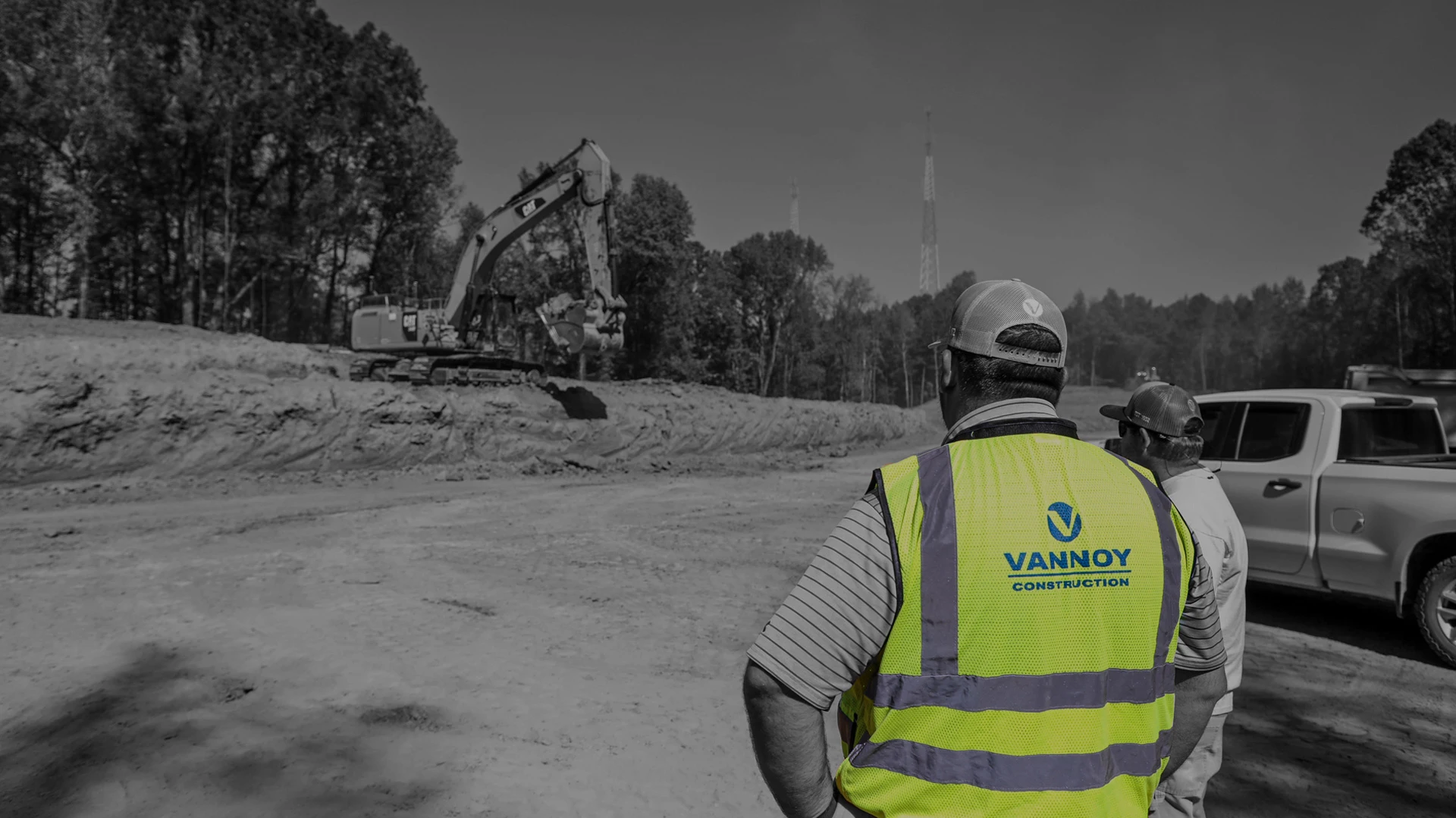 HONOR_2025 A construction worker in a Vannoy Construction vest observes an excavator operating at a dirt worksite with trees in the background.