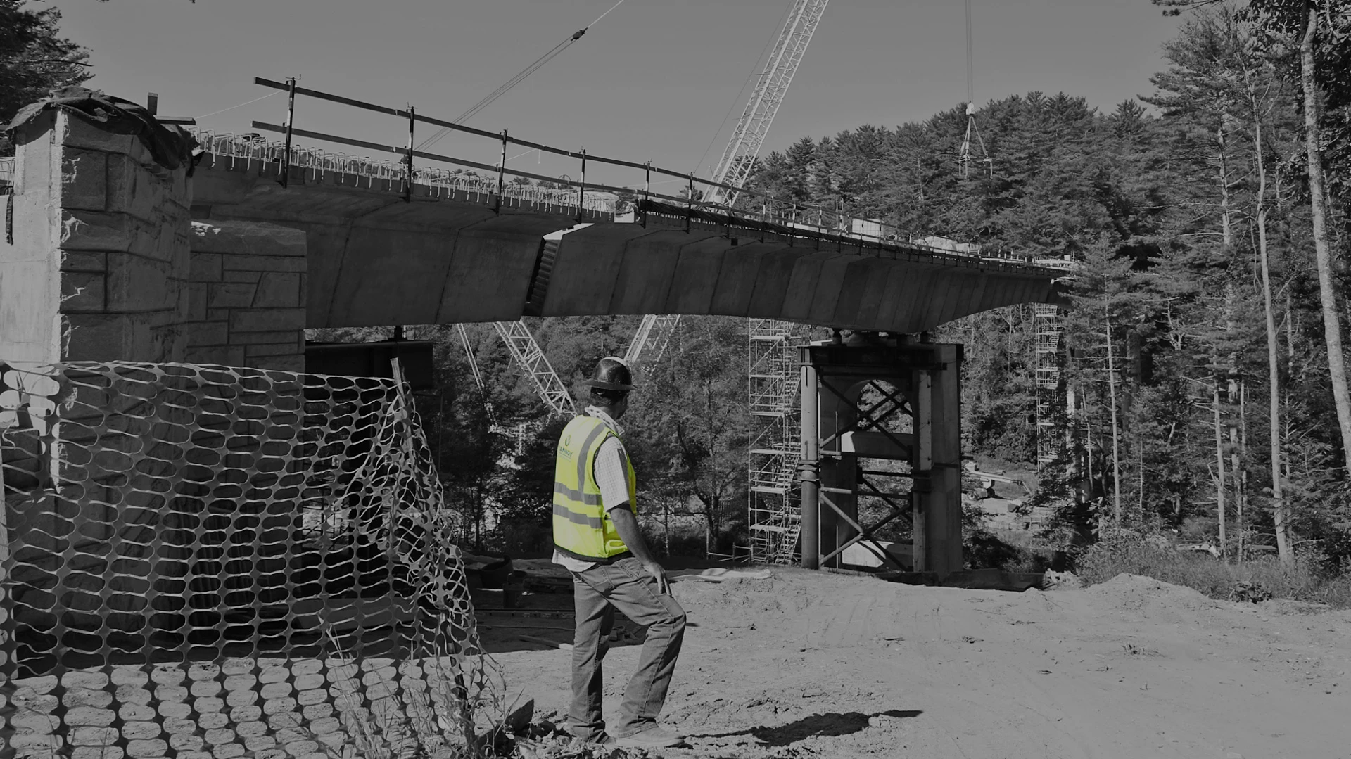 HUM_2025 A construction worker in a safety vest stands near a partially built bridge with cranes and scaffolding in the background.