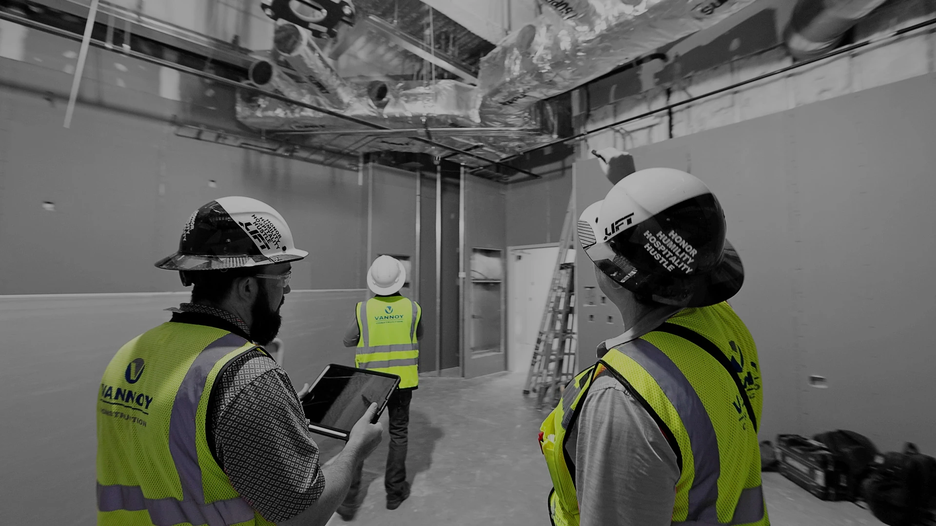 HUSTLE_2025 Three construction workers in safety gear inspect an unfinished building interior; one holds a tablet, and exposed ducts and a ladder are visible.