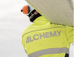Rectangle 5 (2) Person wearing a yellow high-visibility jacket labeled “ALCHEMY” and protective gear, seen from behind, holding a large object on their shoulder.