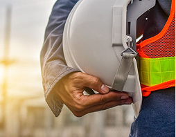 Rectangle 5 (8) Person holding a white safety helmet and wearing a reflective orange safety vest, standing outdoors at a construction site.