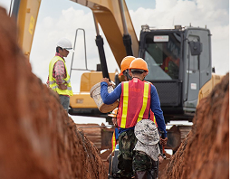 Rectangle 5 Construction workers wearing safety gear work in a trench near a large excavator on a cloudy day.