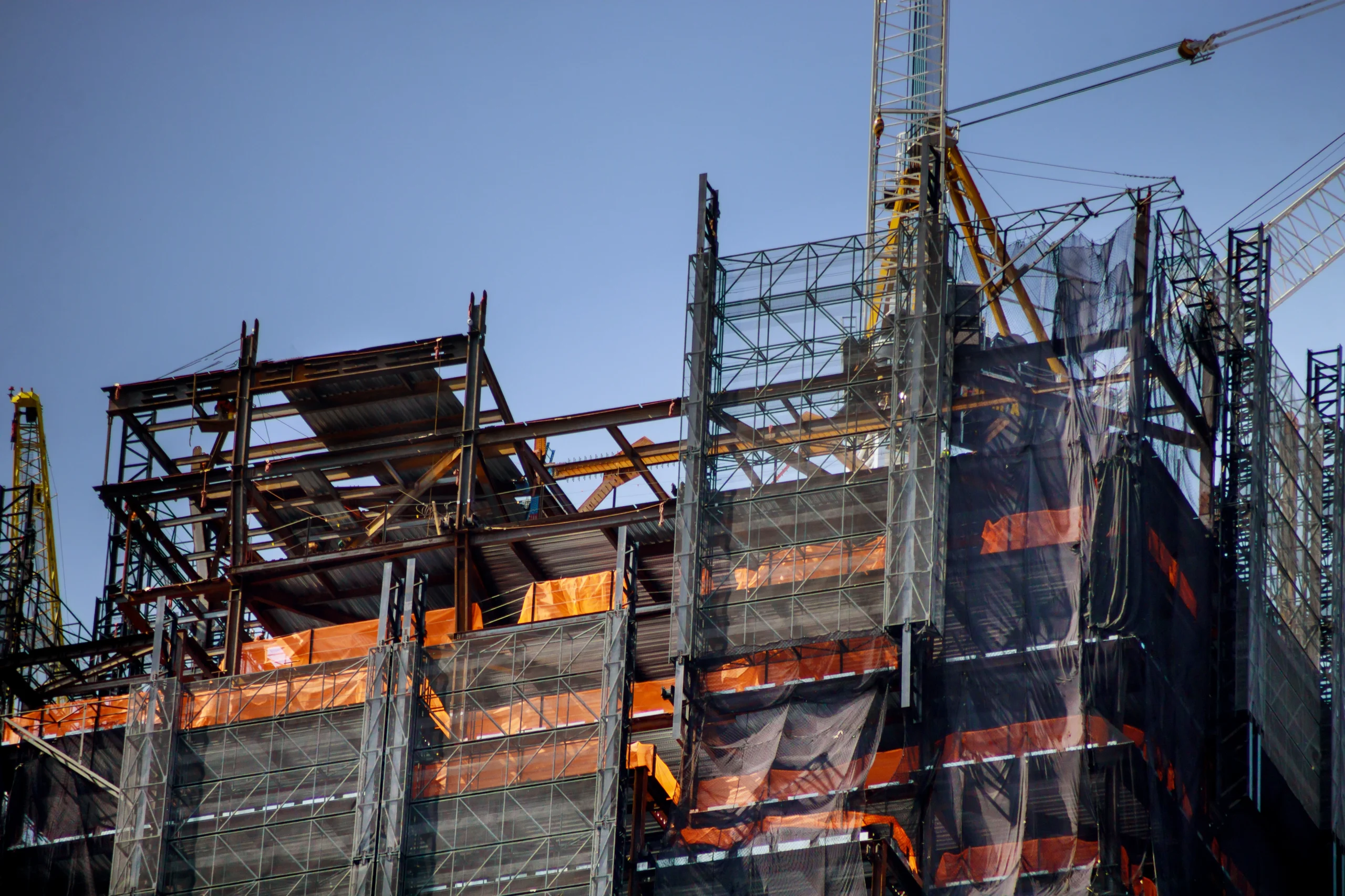 construction-work-site-residential-lots-of-tower-c-2026-03-25-00-37-11-utc Steel framework and scaffolding of a multi-story building under construction, with cranes and safety netting visible against a clear blue sky.