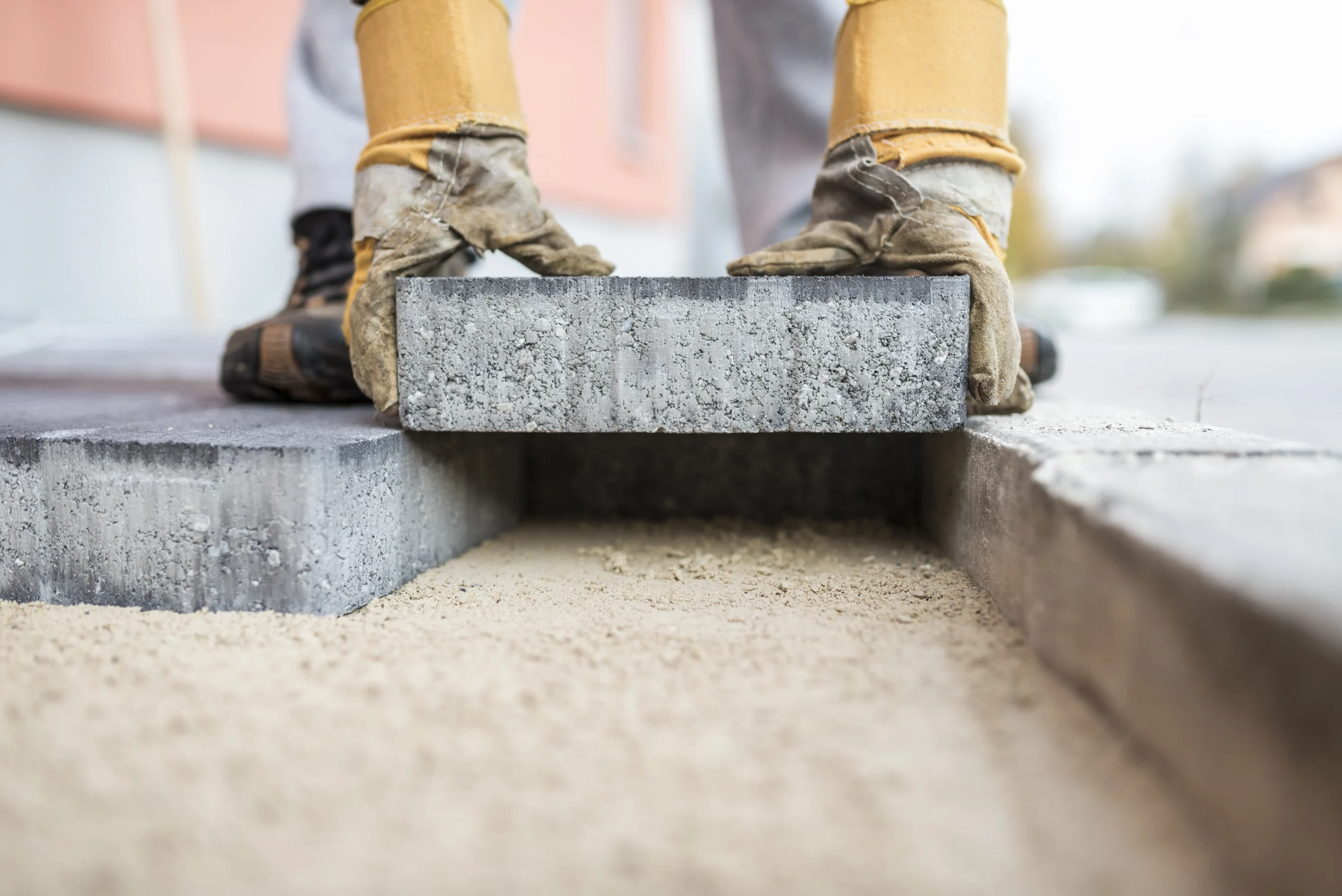 construction-worker-laying-pavers-for-an-outdoor-s-2026-03-19-01-59-02-utc A person wearing gloves places a rectangular concrete paver onto a bed of sand during a paving project.