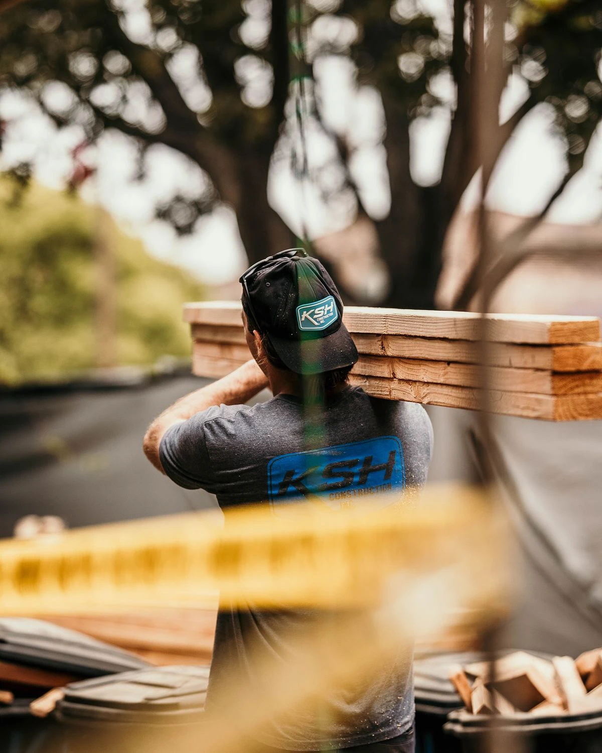 e0a4f74119ab936df9e69a37e108b69d A man in a KSH shirt and cap carries several wooden planks on his shoulder at an outdoor construction site, with trees and caution tape visible in the background.
